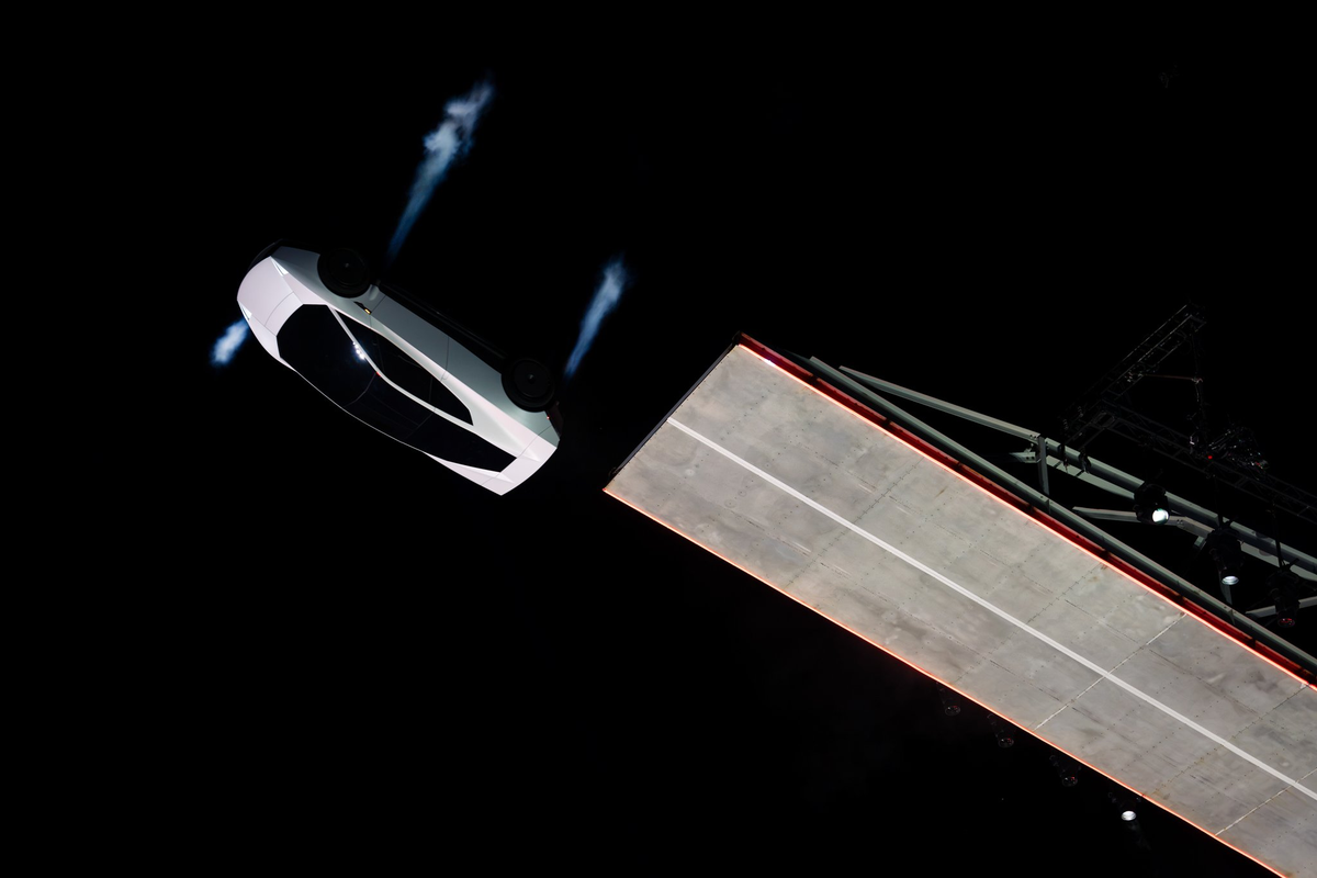 Overhead view of a white Tesla Roadster hovering on four thruster plumes beside a lit ramp at night.